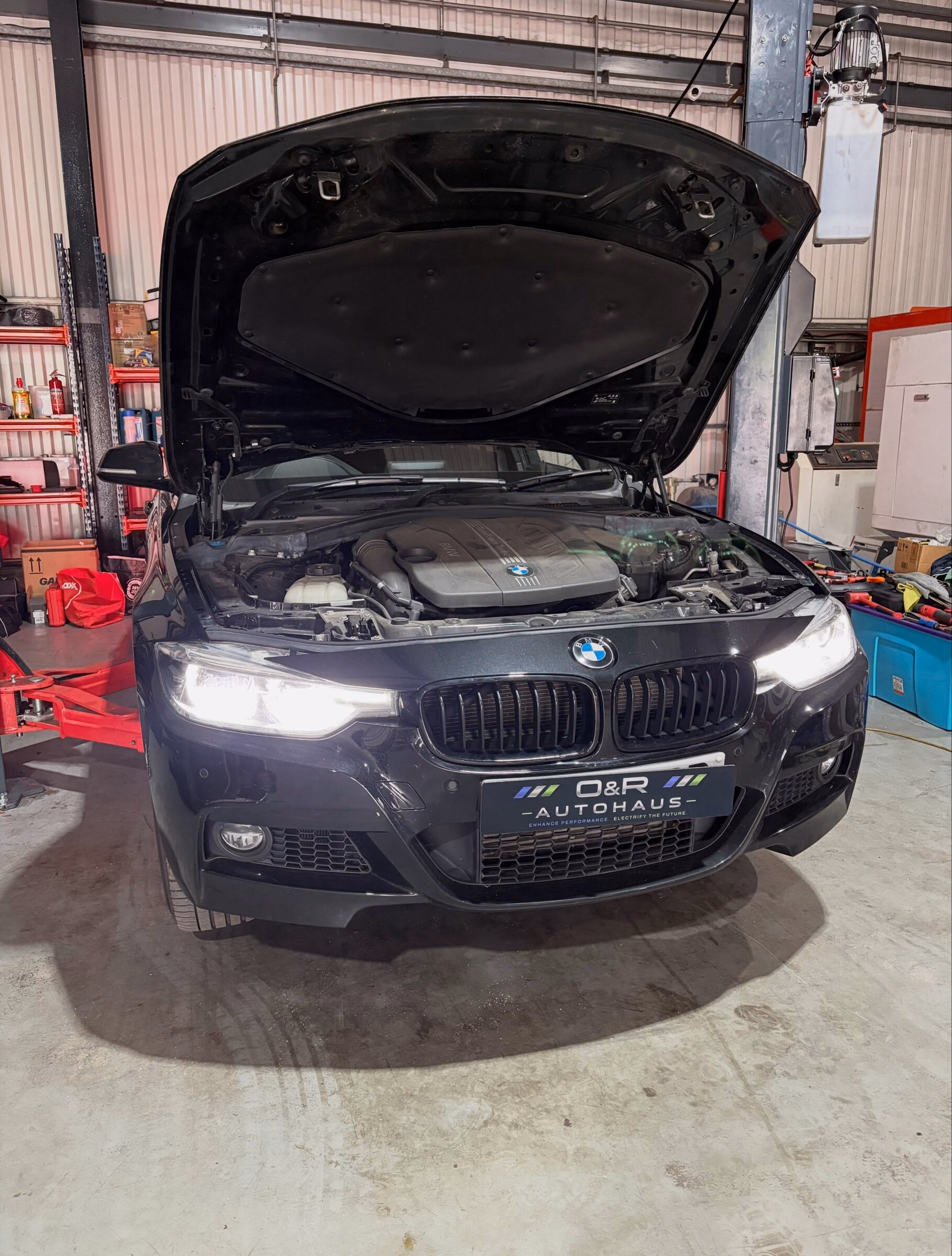 A BMW 3 Series on the bay at O&R Autohaus with its bonnet up and an inspection lamp over the engine