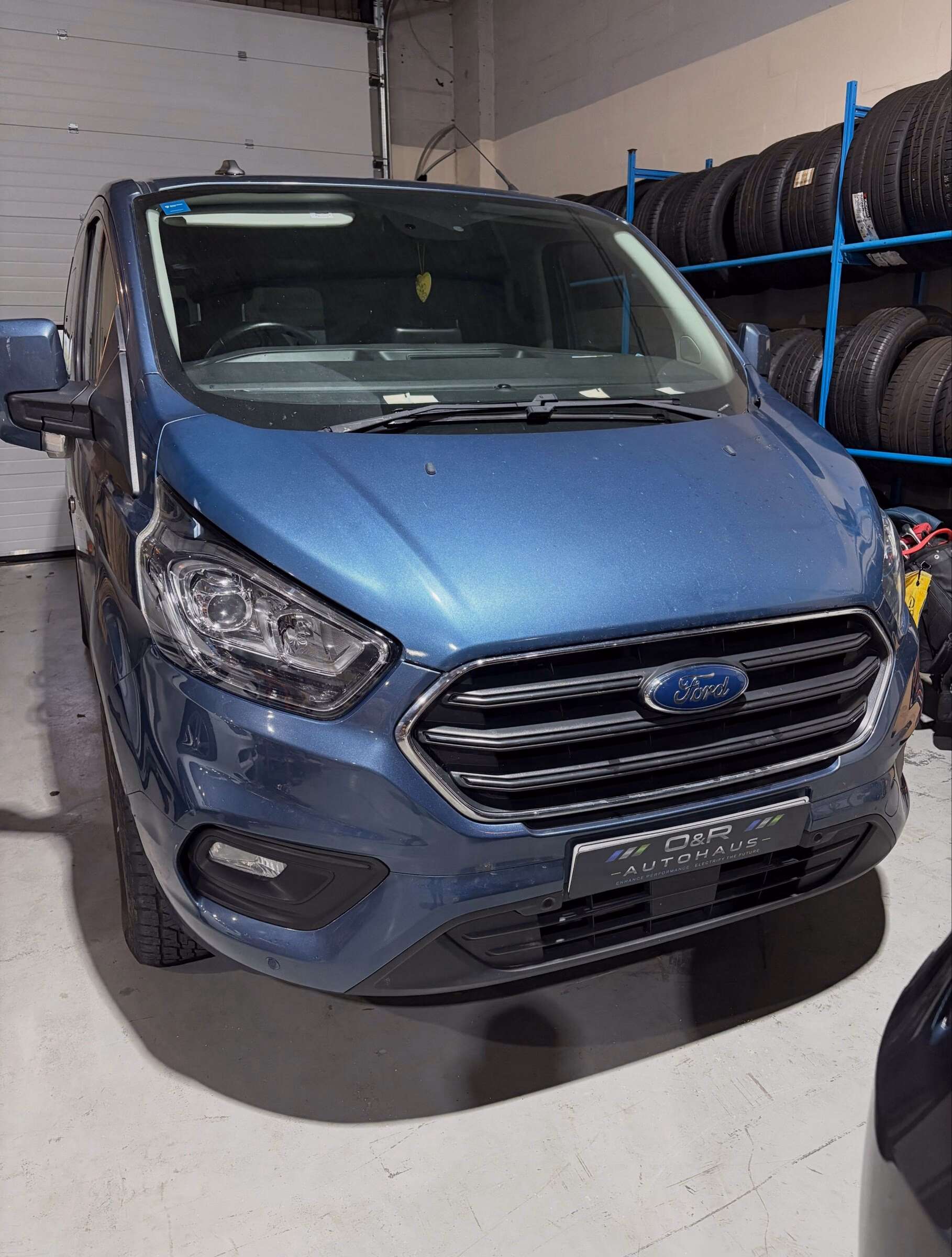 A blue Ford Transit Custom in the workshop at O&R Autohaus with the tyre rack behind it