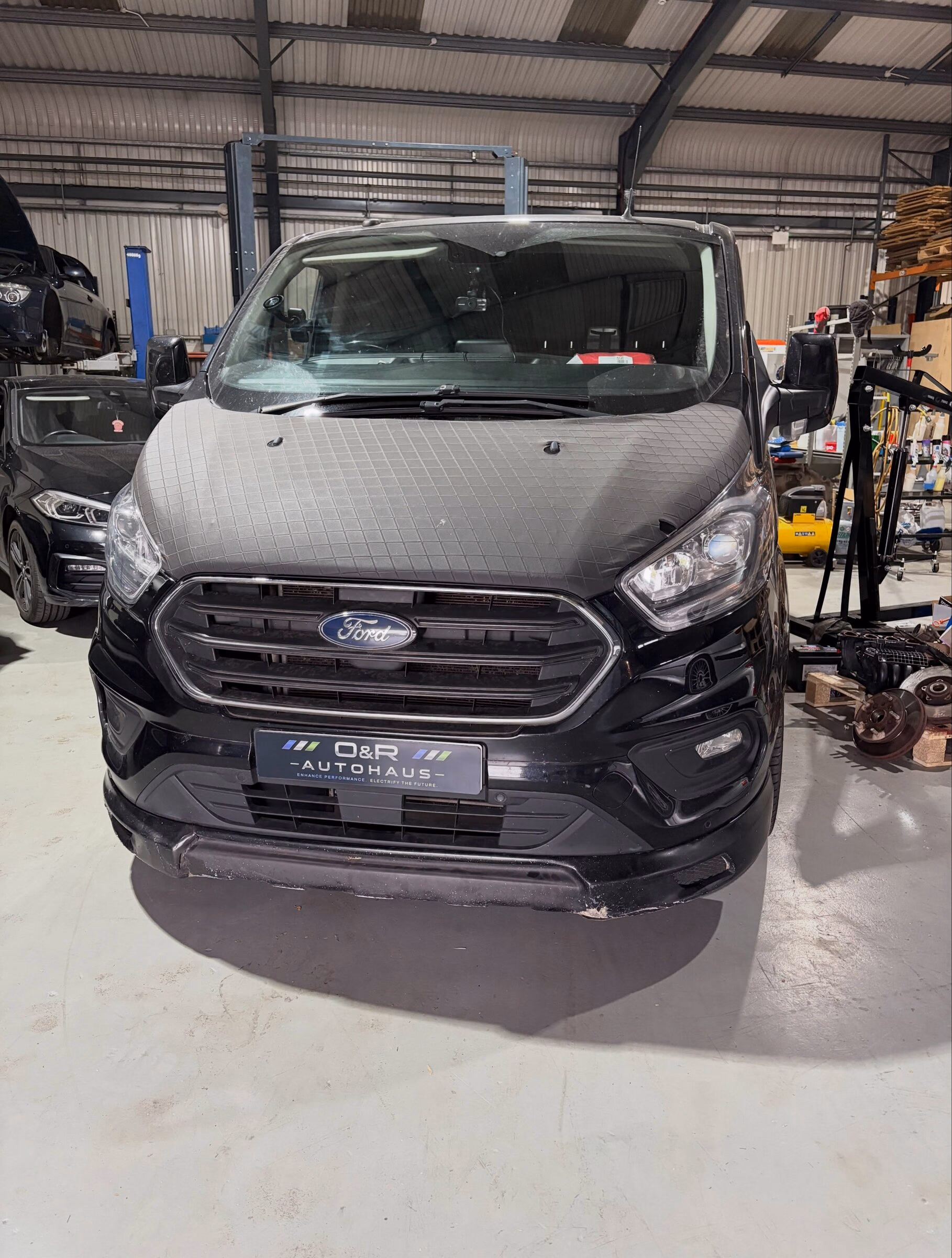 A Ford Transit Custom in the workshop at O&R Autohaus with another vehicle on the ramp behind it
